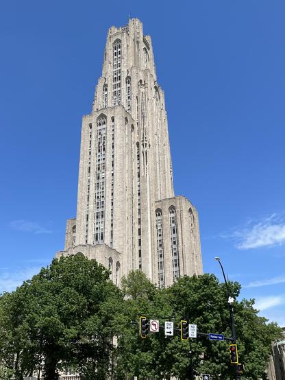 Photo of a tall, gracefully sculpted stone building approximately 36 stories tall, with green leafy trees all around the base. A brilliant blue sky backs the scene.
