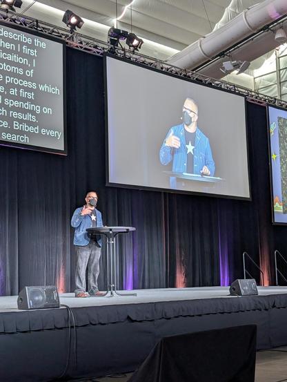 Cory Doctorow is on stage, behind a cocktail table. he's wearing a filtration mask. there's a bigger pic of himself in the background (it's a projection screen)