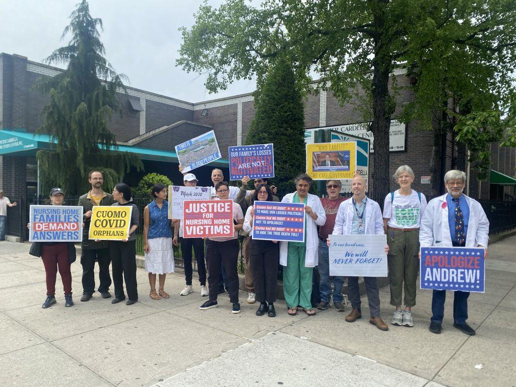 Group of about 15 people, including senior citizens and health care workers, facing the camera as a group, in front of a Senior Center in Queens. They hold signs reading ＂Nursing Home Families Demand Answers＂, ＂Cuomo COVD Orphans＂, ＂Justice for Nursing <br>Home Victims＂, etc. My wife (tall, grey-haired, wearing glasses) sports a tee shirt which reads ＂New York City: Don't Rank Cuomo!＂