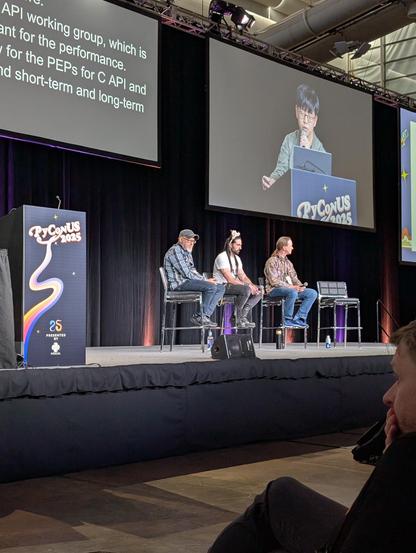 three core developers on stage in bar stools at PyCon US. Pablo, the middle one, is wearing some strange rabbity ear thingies.