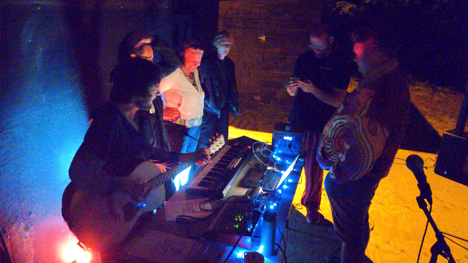 People around a table, singing, playing the guitar and keyboards; in the dark but with colorful lights around.