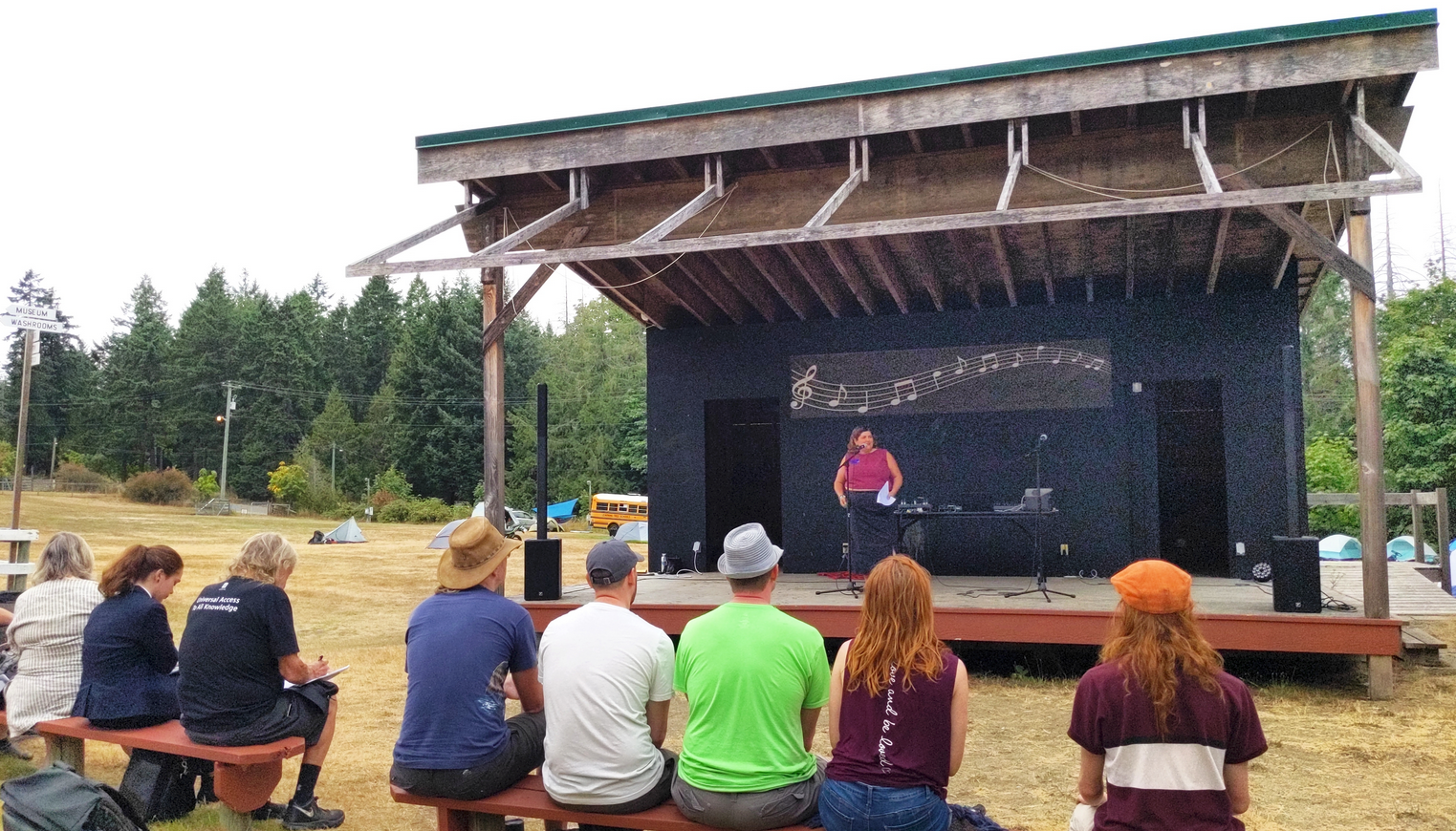 A few people are seated and listening to a woman talking on a stage.