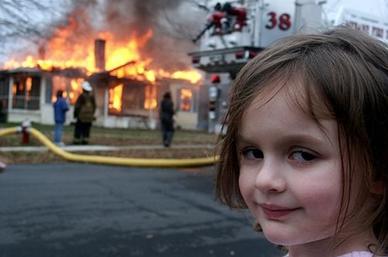A photo. In the background, a house is on fire. In the middle ground you can see firefighters, a hose, and part of a ladder truck. In the foreground, a young child smiles mischievously at the camera.