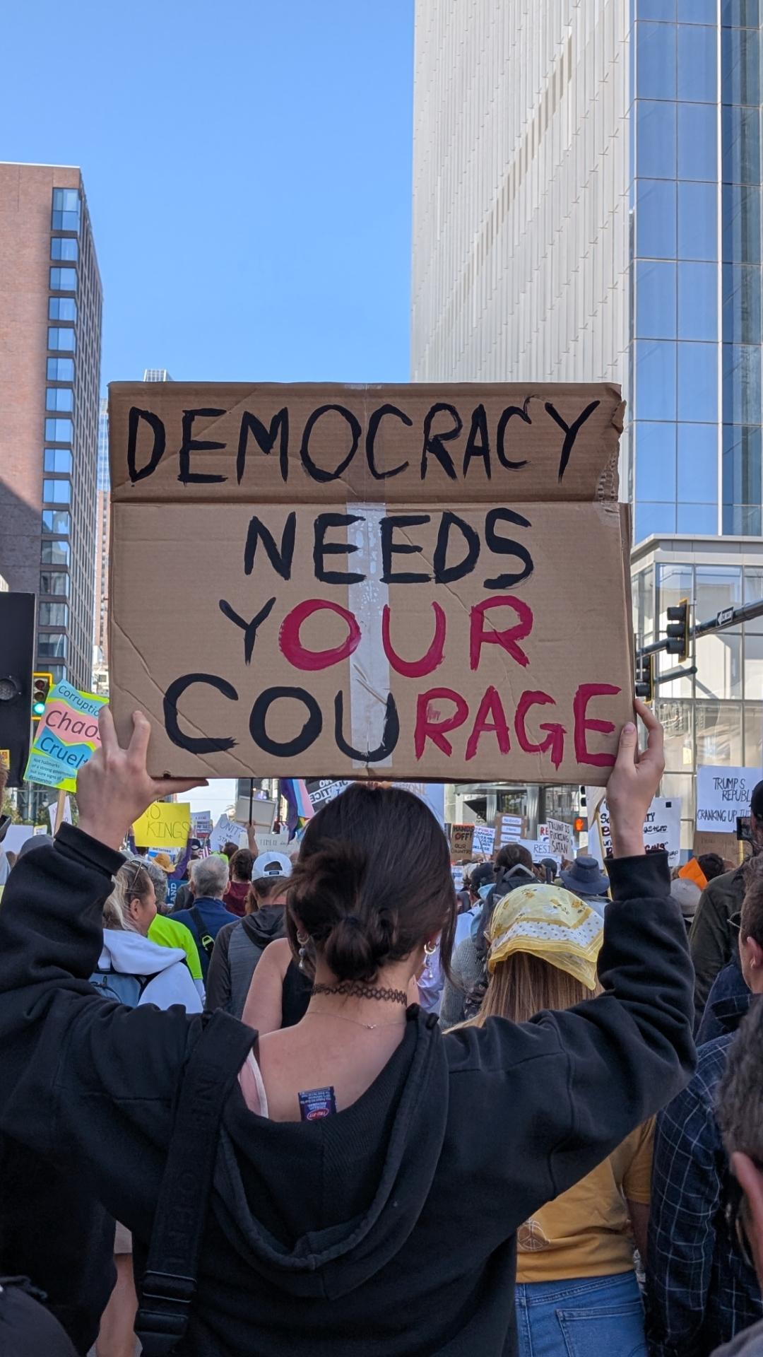 A protester holding a cardboard sign with the words, "Democracy needs your courage." All the letters are in black except a subset in red which makes the sign also read, "Our democracy needs our rage."