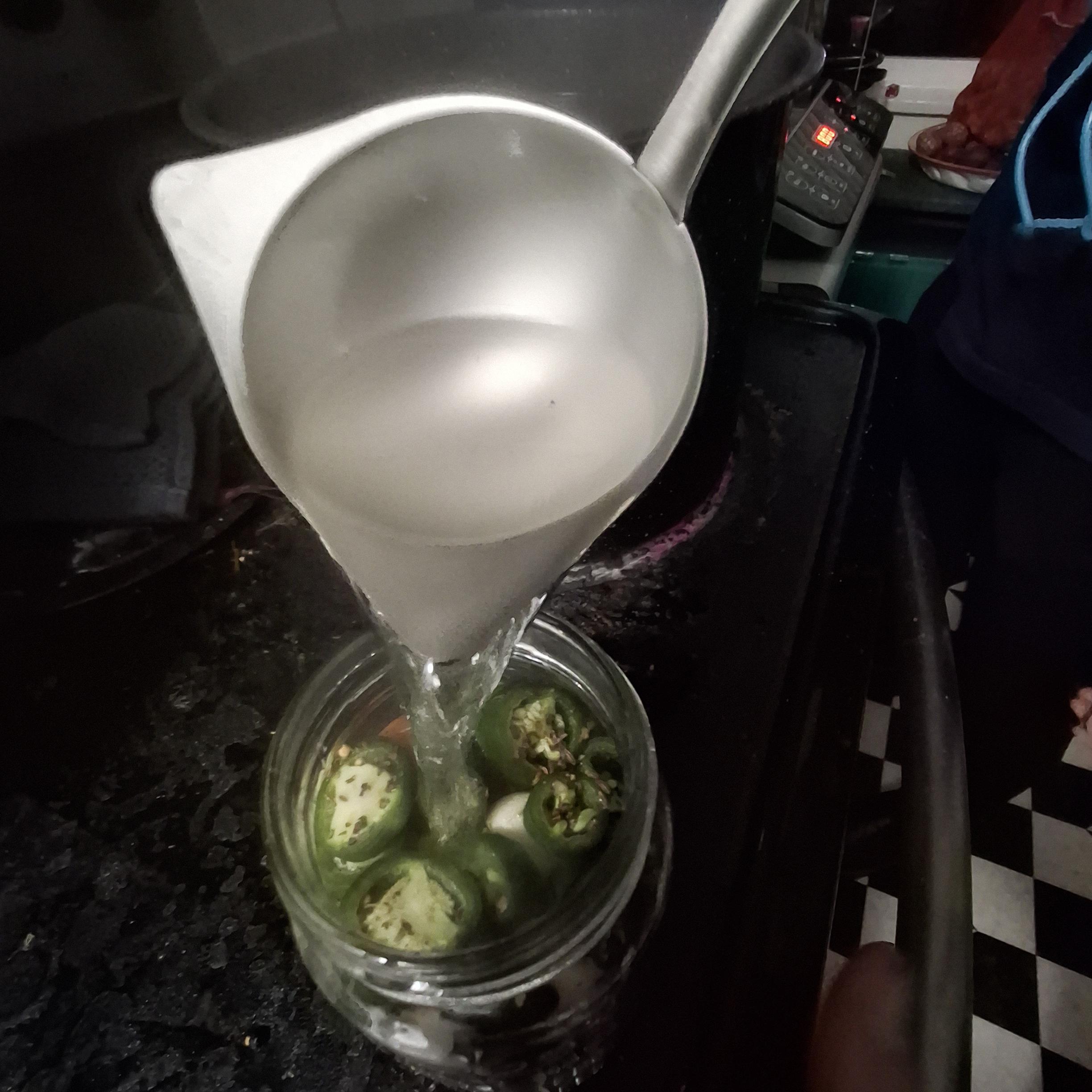 Brine being ladled into a glass jar full of green peppers.