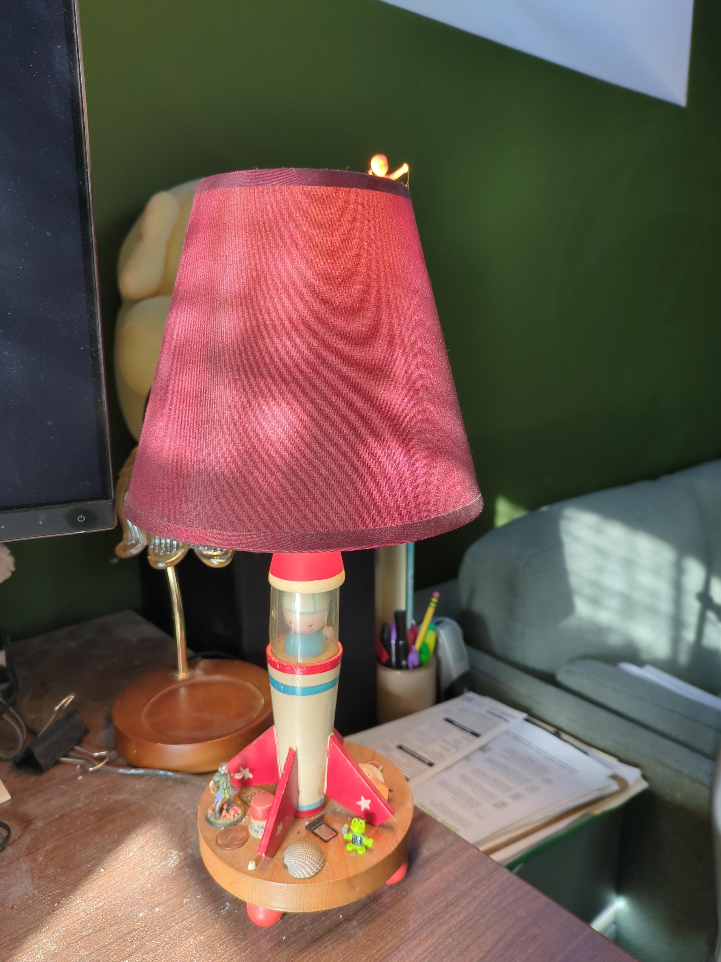 A lamp with a round unpainted wood base sitting on spherical red feet on my desk. The body of the lamp is shaped and painted like a rocket ship, and 2/3 up there is a carving of a little astronaut guy, with a sheet of clear plastic around him to imitate glass. The lampshade is red, not a good match for the red on the rocketship but oh well.

On the base of the lamp between the rocket fins rest several trinkets & doodads, including two seashells, one of my childrens' baby teeth, a custom mini of an old TTRPG character of mine, and a frog that's maybe supposed to be Thor? A plastic king cake baby is stuck on the top edge of the lampshade as though riding it like a merry-go-round

Background includes a different lamp in need of repair, and my Mastodon plushie.