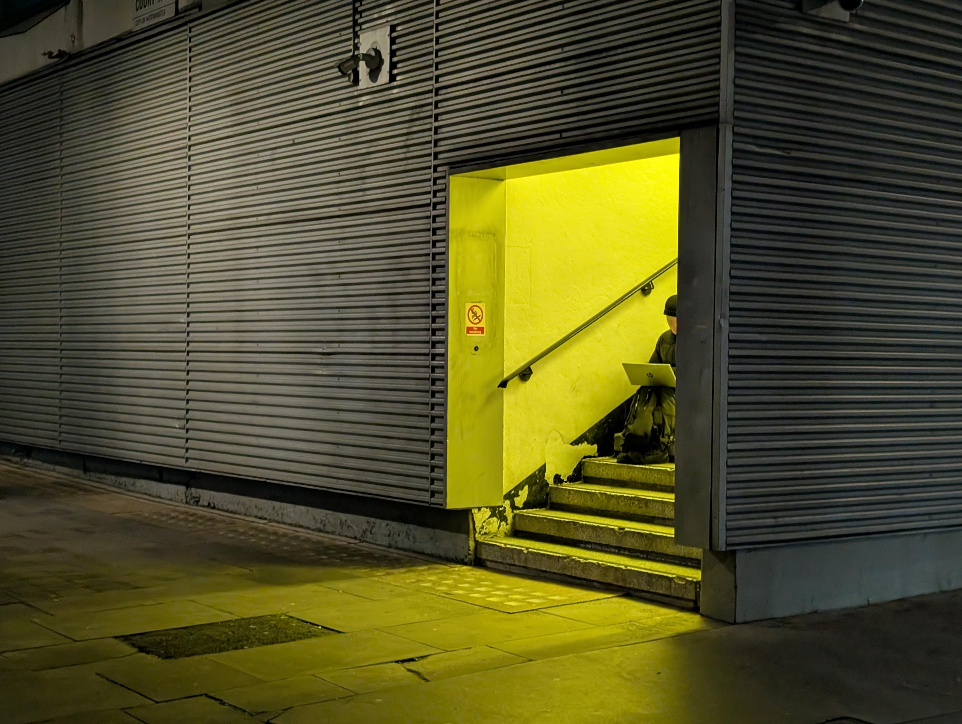 A side entrance to an unmarked building with walls covered in corrugated metal. A bright yellow neon light spills out from the doorway. Inside, someone sits hunched over a computer, working intently on something.