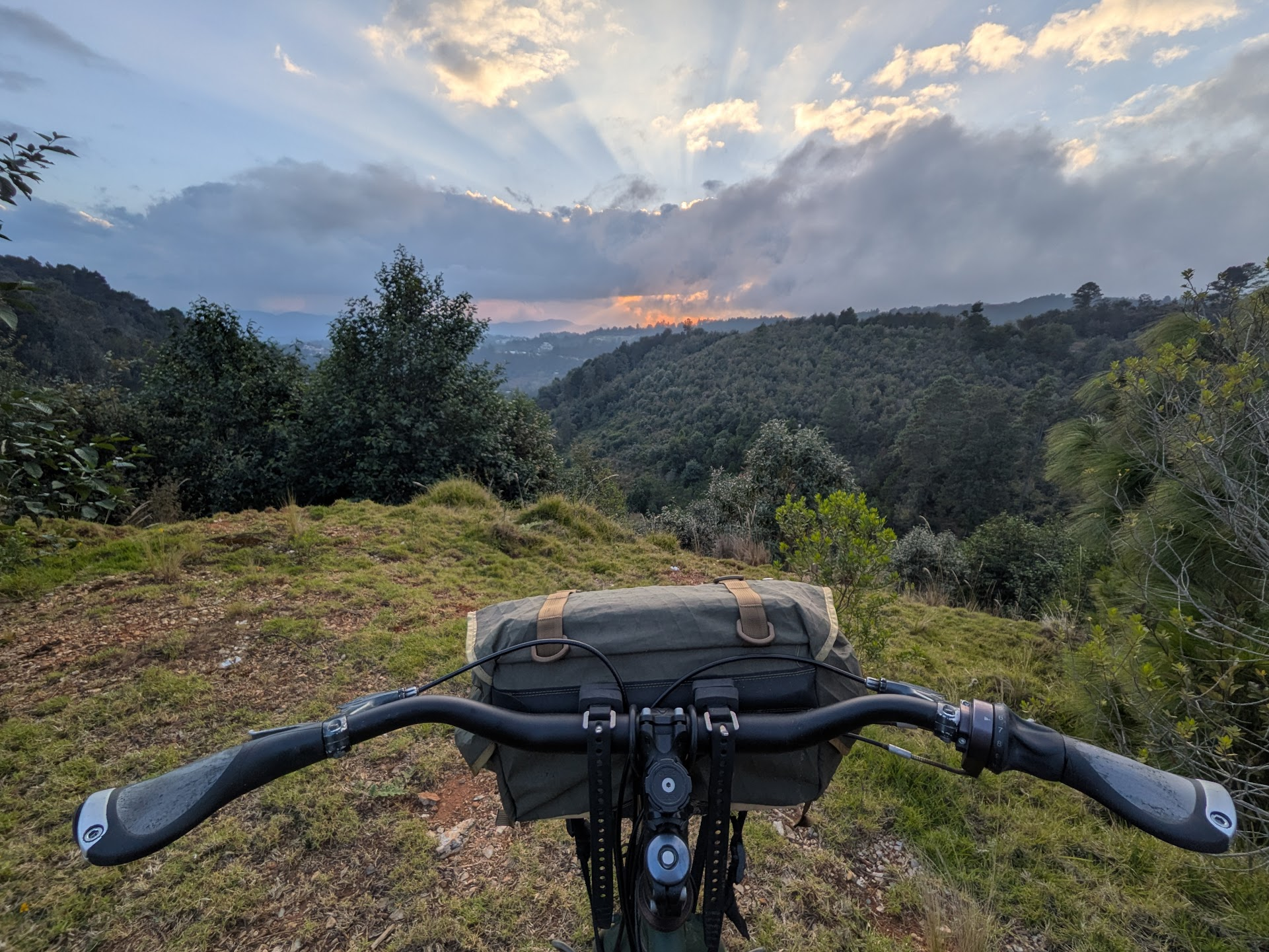 View over bicycle handlebars in the foreground, looking out across a green, forested valley. The setting sun is hidden behind a bank of clouds, with god-rays streaking the sky above and below.