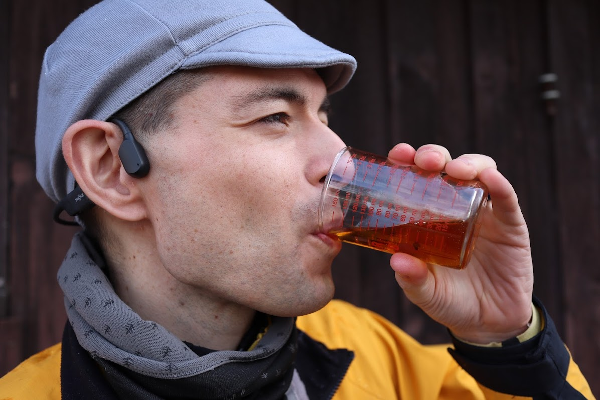 Photograph of a person with a gray wool hat and scarf drinking a clear glass measuring cup of maple syrup.