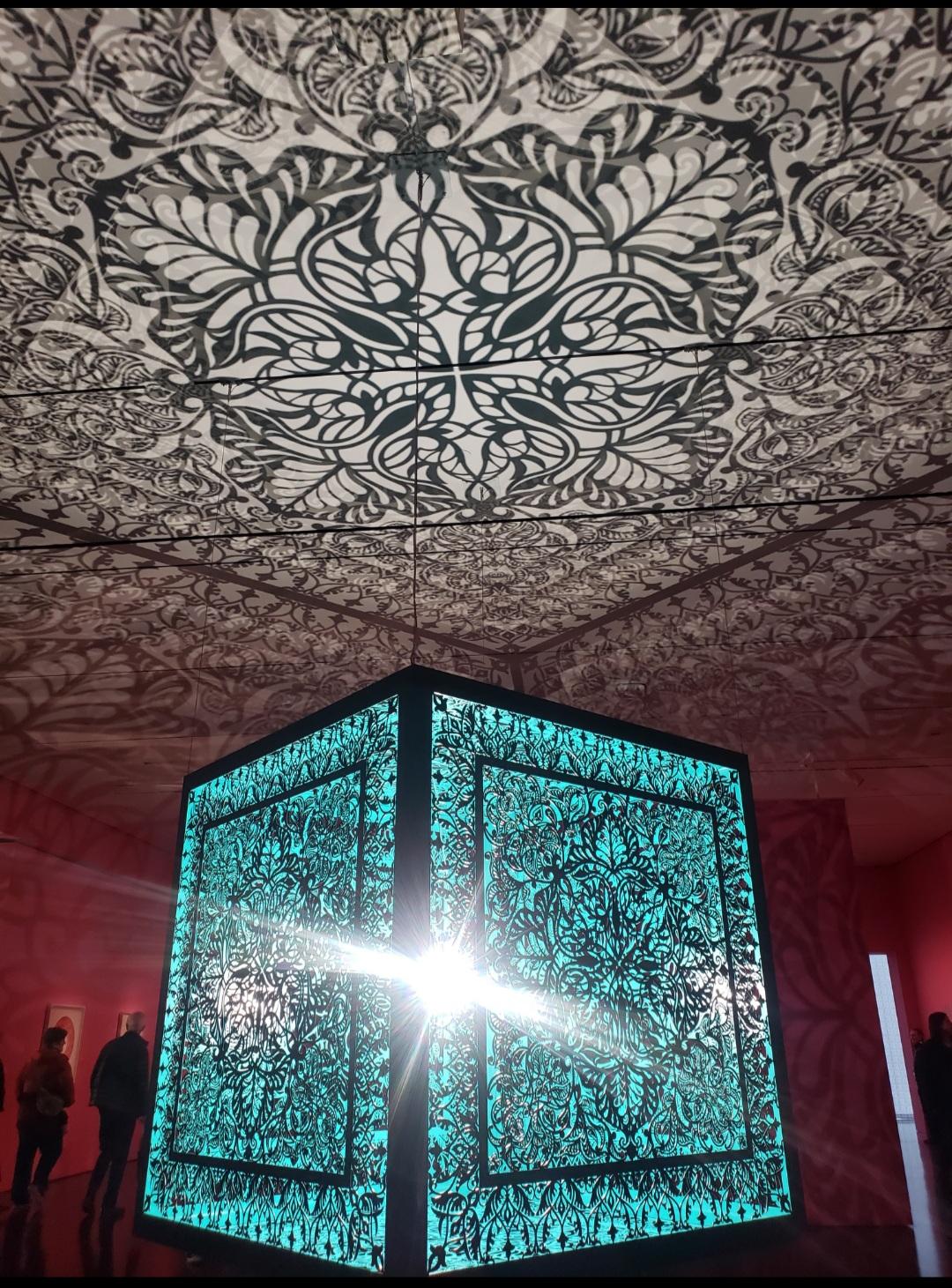 A very large metal cube, with the sides laser cut with intricate patterns, is hanging from the ceiling in an otherwise empty museum room. The main light in the room comes from inside the cube, which reflects the shades of the patterns all around the room, in the ceiling and walls. The cube itself looks blue, the ceiling white and the walls reddish. A couple of museum goers stand around.