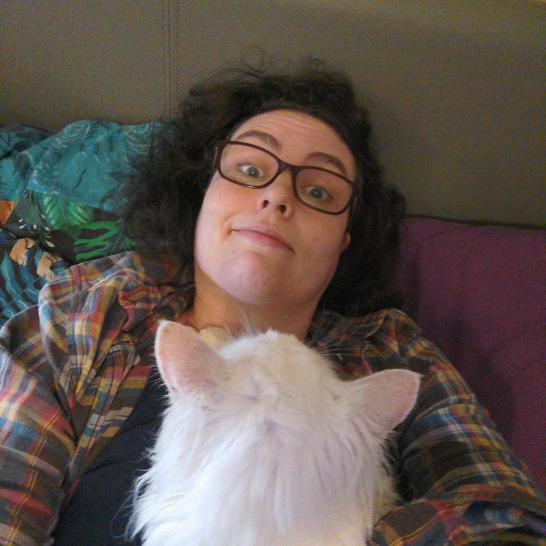 Me, a white woman with dark curly hair, glasses and an expression of resignation to not moving until I am released, lying on a bed with a fluffy white cat on my chest. My head is tilted away from the tuna breath, and his little paws are on my windpipe in a manner that is either affectionate or threatening, it's sometimes hard to tell with cats