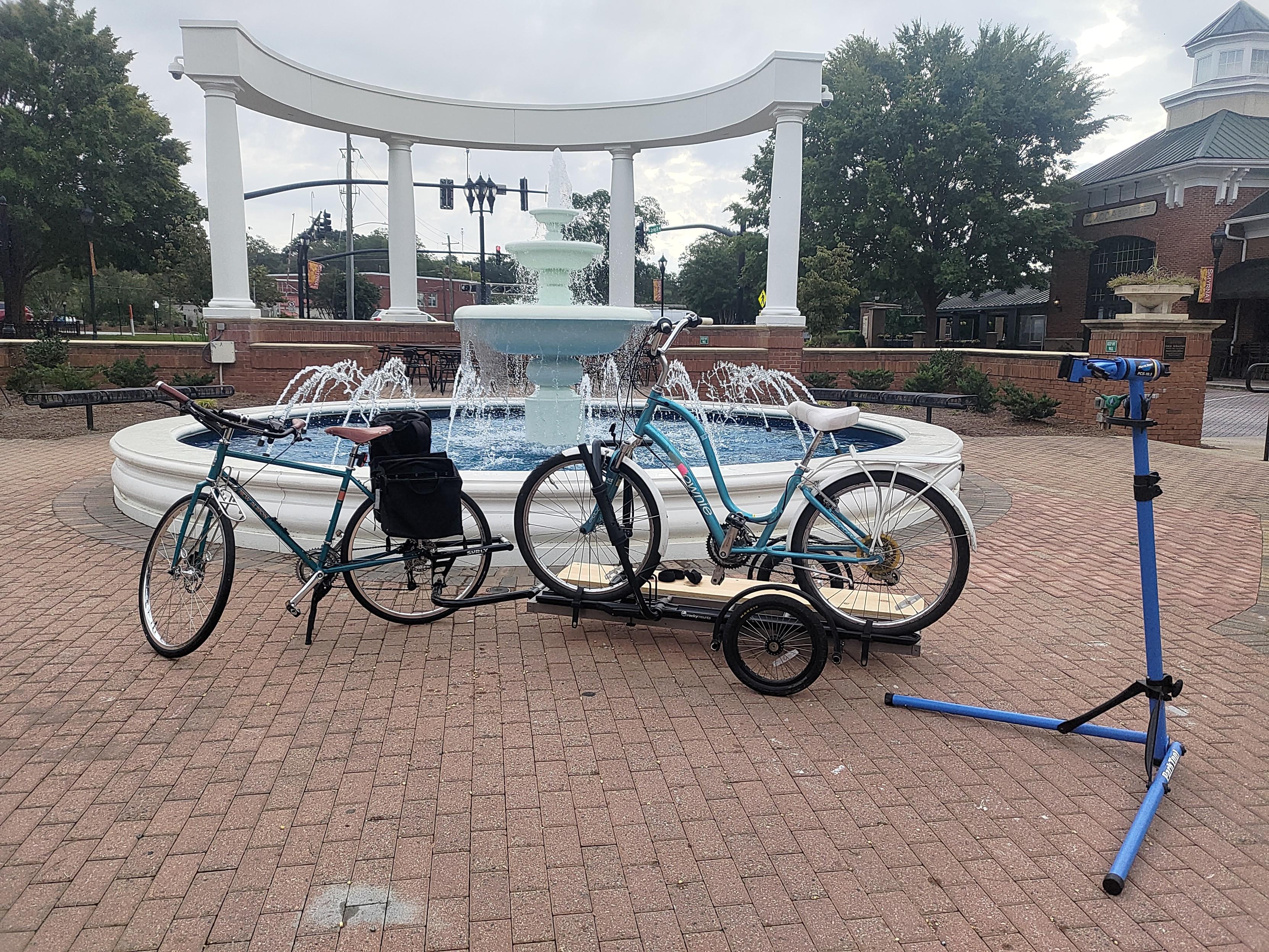 A blue bicycle in a brick town-square towing a trailer on which another blue bicycle (this one a step-thru) is mounted to a bike stand. To the right is a bicycle repair stand that is set up but has presumably also been hauled on the same trailer. In the background is a water fountain and several decorative pillars.