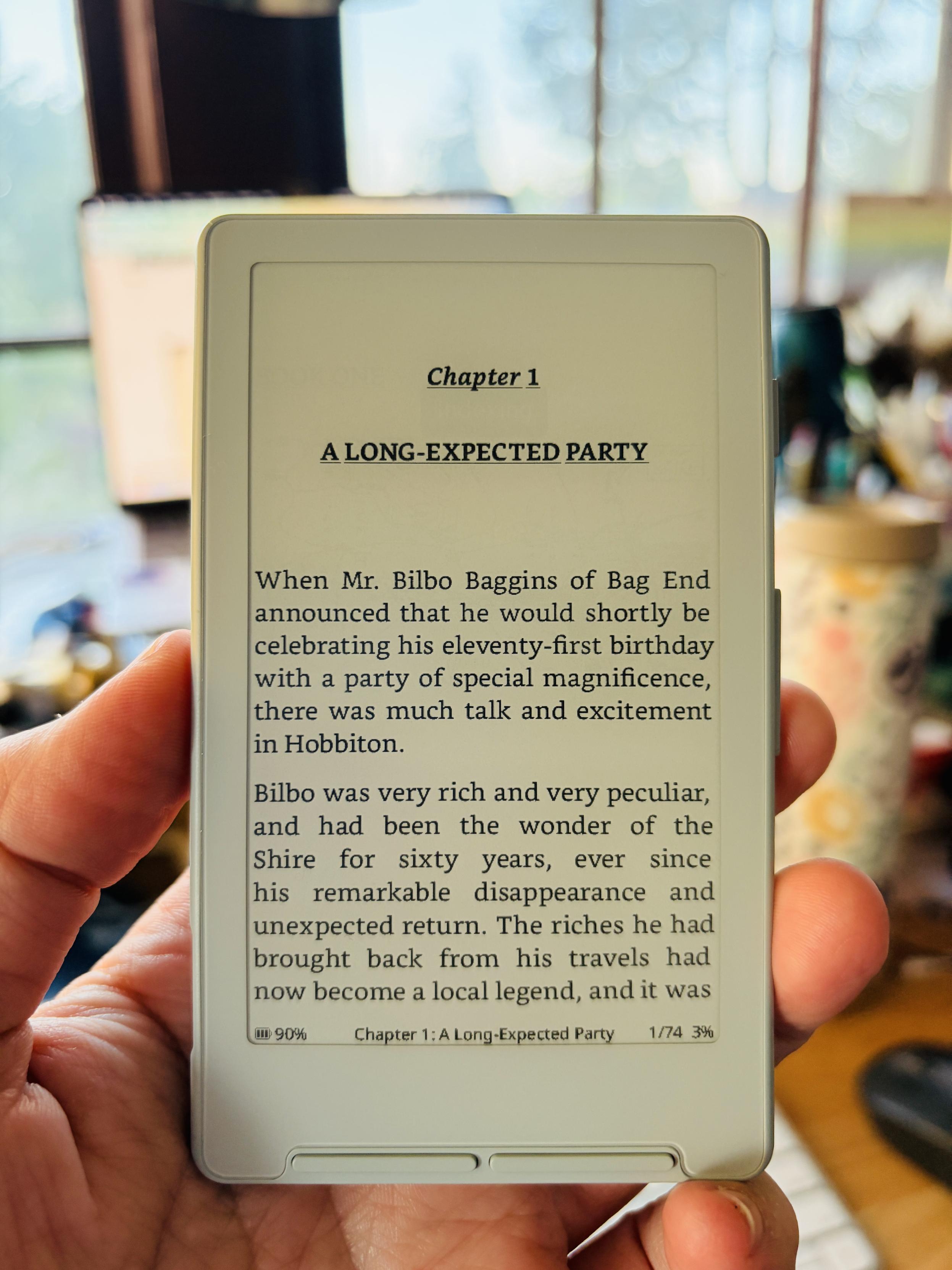 A hand holding a very small ereader open to the first page of The Fellowship of the Ring.