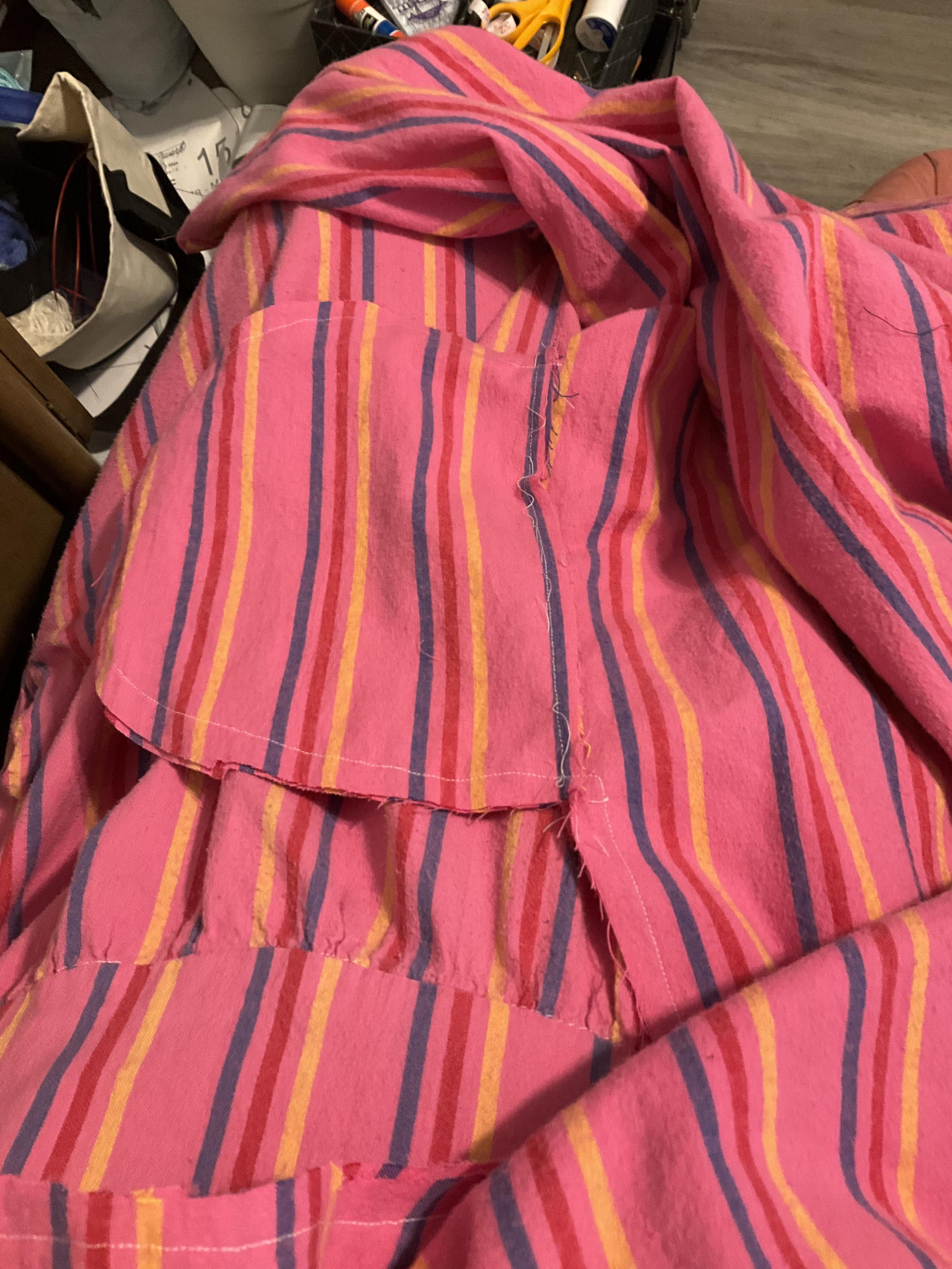 A mass of pink fabric with yellow blue and red stripes. It’s a top down viewof my lap, so you can see the waistband of the dress i am wearing and the pocket and sleeve of the dress i am sewing