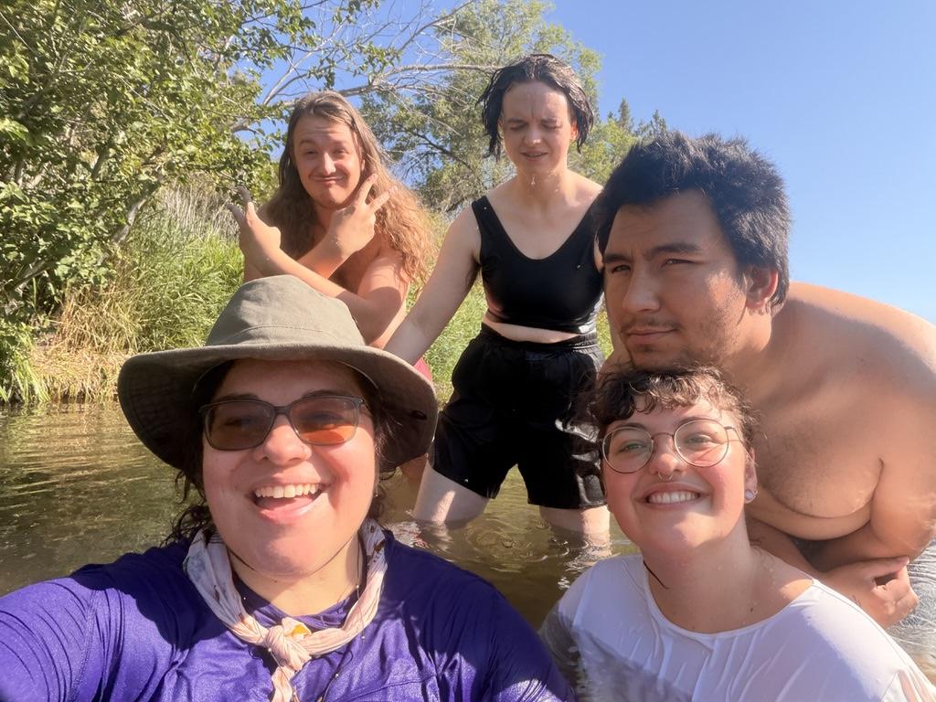 Five wet people standing and sitting in a river, taking a group selfie.
