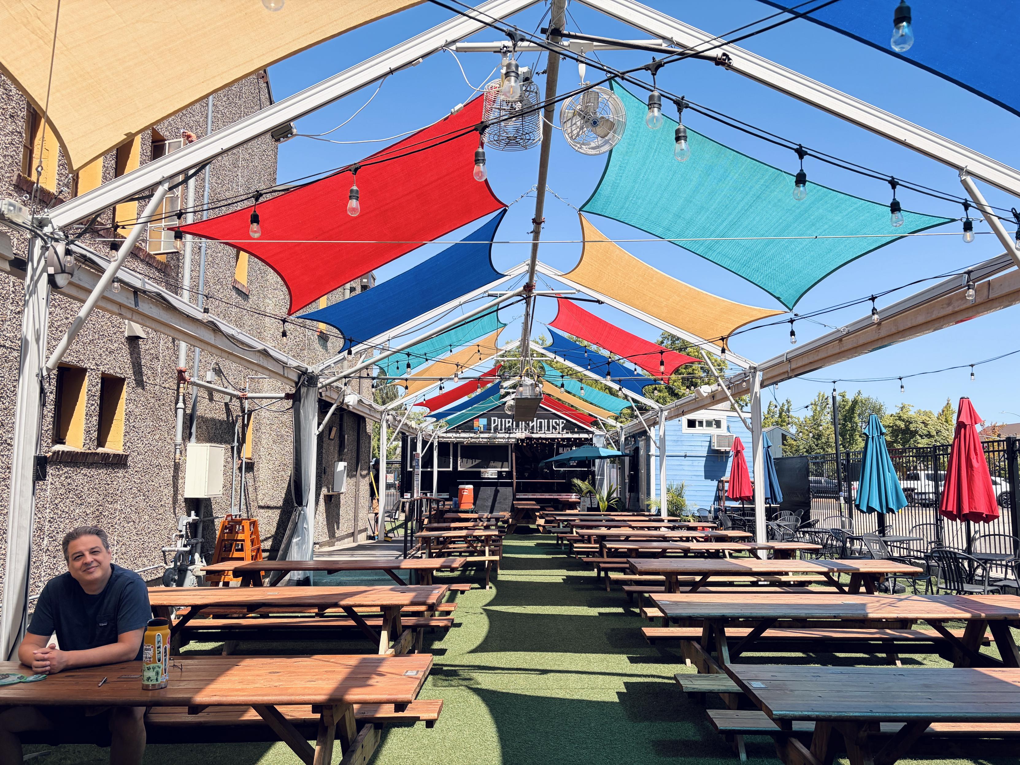 Devastatingly handsome man sits at a picnic table, amongst many, in the back outdoor seating area of a chuch-turned-food court. Along the "ceiling" of the area are stretched pieces of canvas for shade, in various colors.